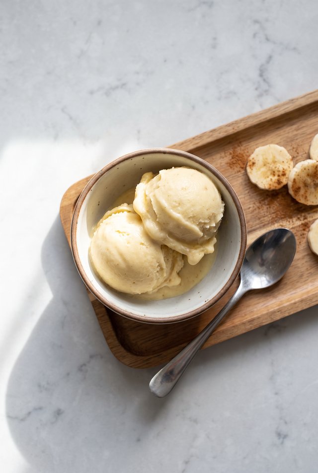 Two scoops of creamy banana nice cream in a bowl on a wooden tray with cinnamon-dusted banana coins beside it