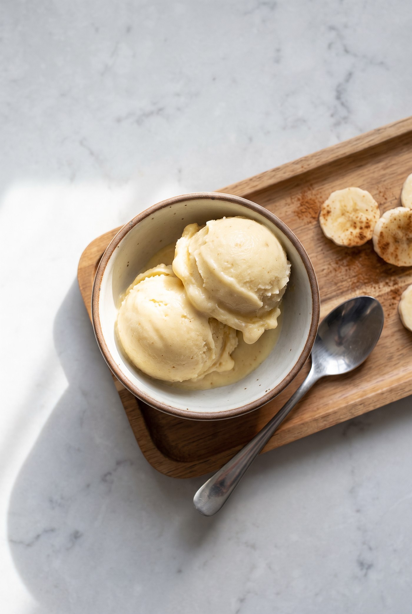 Two scoops of creamy banana nice cream in a bowl on a wooden tray with cinnamon-dusted banana coins beside it