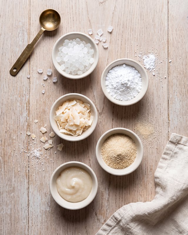 Five small ceramic bowls of pale baking ingredients on a wooden counter, granulated and powdered sweetener, coconut flakes, almond flour and a thick syrup, with a brass measuring spoon alongside