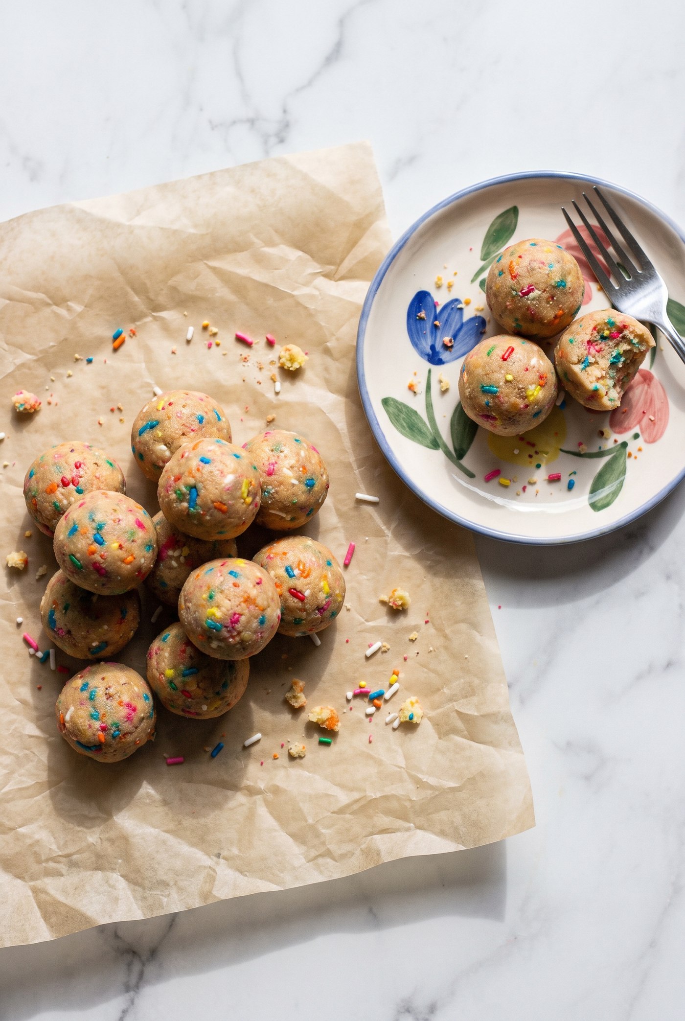 Pile of sprinkle-studded birthday cake protein cake balls on parchment paper next to a floral plate with a bitten one on a fork