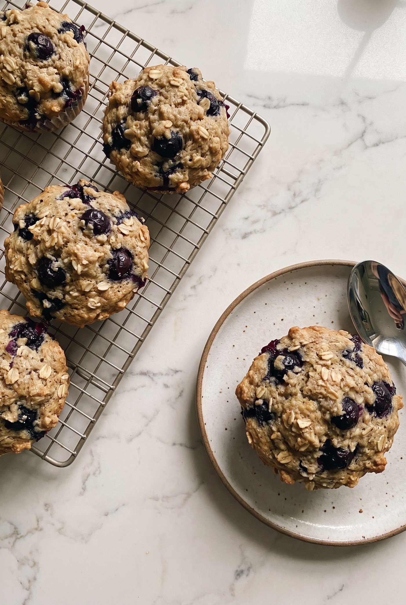 Blueberry oat muffins cooling on a wire rack with fresh blueberries scattered nearby, one muffin plated with a spoon