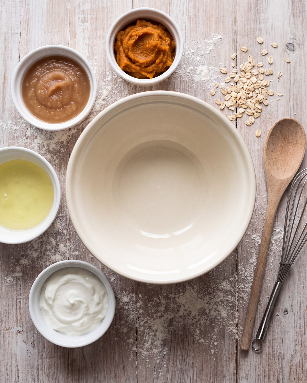 Small bowls of applesauce, pumpkin puree, egg whites and yogurt around a mixing bowl on a floured surface